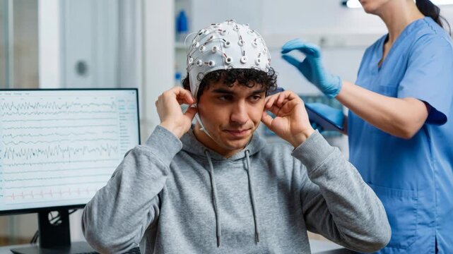 Young man wearing wearable EEG cap and biosensor for brain monitoring in clinical neurology clinic with technician observing EEG data on screen