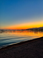 Fototapeta premium Evening sunset at Issyk Kul Lake in Kyrgyzstan. Long illuminated pier stretching into calm water with mountains on horizon.