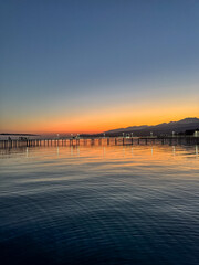 Fototapeta premium Evening sunset at Issyk Kul Lake in Kyrgyzstan. Long illuminated pier stretching into calm water with mountains on horizon.
