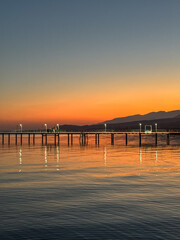 Fototapeta premium Evening sunset at Issyk Kul Lake in Kyrgyzstan. Long illuminated pier stretching into calm water with mountains on horizon.