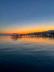 Fototapeta premium Evening sunset at Issyk Kul Lake in Kyrgyzstan. Long illuminated pier stretching into calm water with mountains on horizon.