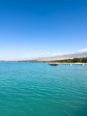 Fototapeta premium Issyk Kul Lake in Kyrgyzstan on a sunny day. Turquoise water, long pier and Tien Shan mountains on the horizon.