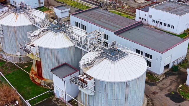 Storage tanks stand in rows next to large buildings in an industrial zone. There are green areas and paved paths around the structures during daylight.