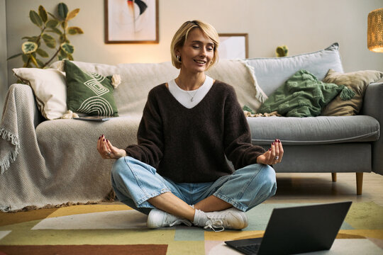 Caucasian woman sitting cross legged on floor meditating in front of open laptop, practicing mindfulness or relaxation exercise in home setting related to femtech health