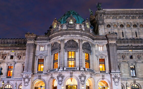 Night view of the illuminated historic architecture and ornate facade of the Palais Garnier opera house in Paris, France.