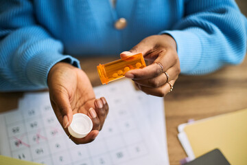 Black woman opening prescription pill bottle, holding medication in hand, sitting at desk with calendar and documents, illustrating femtech health management and daily medication routine © DragonImages