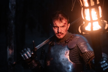 Obraz premium A knight in full armor, sword and torch in hand, walks through a gloomy night forest. A medieval warrior, bloodied and scarred, hunts and searches in the darkness.