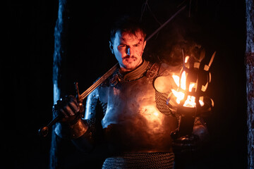 Obraz premium A knight in full armor, sword and torch in hand, walks through a gloomy night forest. A medieval warrior, bloodied and scarred, hunts and searches in the darkness.
