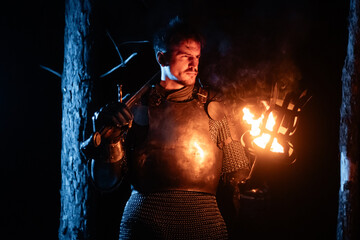 Obraz premium A knight in full armor, sword and torch in hand, walks through a gloomy night forest. A medieval warrior, bloodied and scarred, hunts and searches in the darkness.