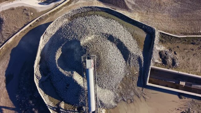A winding path leads to a towering mound of stones in an industrial site, with machinery nearby. Clear skies provide a backdrop as the sun illuminates the rugged textures of the stones.