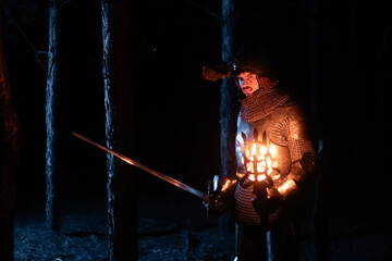 Obraz premium A knight in full armor, sword and torch in hand, walks through a gloomy night forest. A medieval warrior, bloodied and scarred, hunts and searches in the darkness.