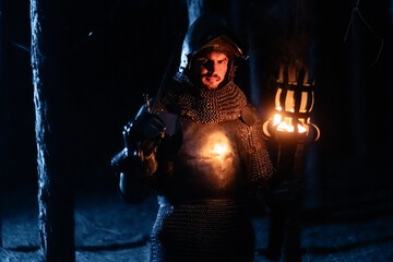 Obraz premium A knight in full armor, sword and torch in hand, walks through a gloomy night forest. A medieval warrior, bloodied and scarred, hunts and searches in the darkness.