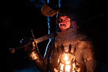 Obraz premium A knight in full armor, sword and torch in hand, walks through a gloomy night forest. A medieval warrior, bloodied and scarred, hunts and searches in the darkness.