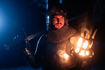 Obraz premium A knight in full armor, sword and torch in hand, walks through a gloomy night forest. A medieval warrior, bloodied and scarred, hunts and searches in the darkness.