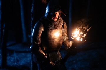Obraz premium A knight in full armor, sword and torch in hand, walks through a gloomy night forest. A medieval warrior, bloodied and scarred, hunts and searches in the darkness.