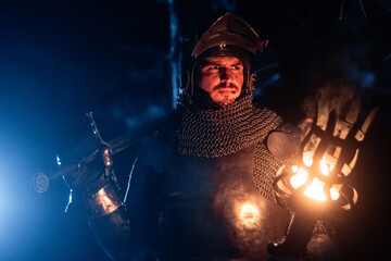 Obraz premium A knight in full armor, sword and torch in hand, walks through a gloomy night forest. A medieval warrior, bloodied and scarred, hunts and searches in the darkness.