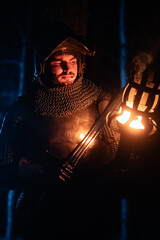 Obraz premium A knight in full armor, sword and torch in hand, walks through a gloomy night forest. A medieval warrior, bloodied and scarred, hunts and searches in the darkness.
