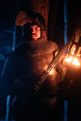 Obraz premium A knight in full armor, sword and torch in hand, walks through a gloomy night forest. A medieval warrior, bloodied and scarred, hunts and searches in the darkness.