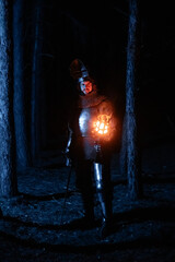 Obraz premium A knight in full armor, sword and torch in hand, walks through a gloomy night forest. A medieval warrior, bloodied and scarred, hunts and searches in the darkness.