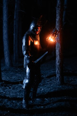 Obraz premium A knight in full armor, sword and torch in hand, walks through a gloomy night forest. A medieval warrior, bloodied and scarred, hunts and searches in the darkness.