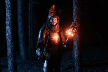 Obraz premium A knight in full armor, sword and torch in hand, walks through a gloomy night forest. A medieval warrior, bloodied and scarred, hunts and searches in the darkness.