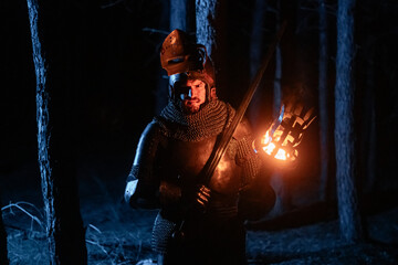 Obraz premium A knight in full armor, sword and torch in hand, walks through a gloomy night forest. A medieval warrior, bloodied and scarred, hunts and searches in the darkness.