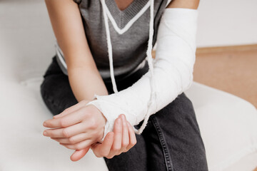 Obraz premium Close up of woman broken arm in white plaster cast gypsum orthopedic sling immobilization sitting on sofa at home during fracture injury recovery rehabilitation treatment showing patient daily life