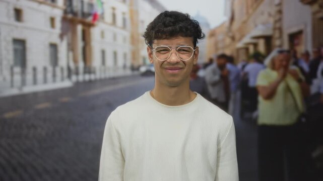 Man wearing glasses and white sweater squints at the bright sun on a crowded urban street; curiosity.
