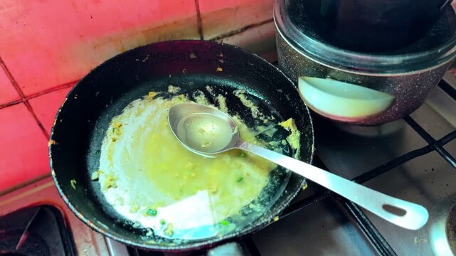 Close-up of a greasy frying pan with oily sauce residue and a metal ladle on a gas stove, beside a covered pot and stained tiled backsplash in a home kitchen.