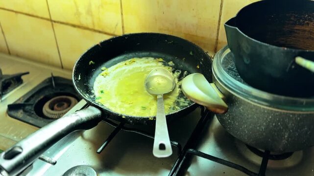 Close-up of gas stove burners with frying pan holding oily sauce residue, ladle and spoon, plus covered pot against a stained tiled backsplash in a home kitchen.