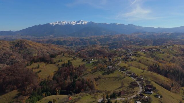 Breathtaking Autumn Village and Hillcrest Road in Pestera under Snow Capped Bucegi Mountains Aerial Descending Shot 4K Graded
