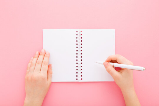 Little child girl hand holding pen and writing on white copybook on light pink table background. Pastel color. Closeup. Empty place for text. Flat lay. Point of view shot. Top down view.
