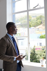 Fototapeta premium Black man, phone and thinking at window in office with texting, contact or feedback at real estate agency. African person, realtor or property developer with smartphone, perspective or chat on web