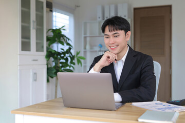 Fototapeta premium Young Asian businessman in a suit working on a laptop, smiling. He is achieving success and online business goals