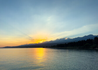 Obraz premium Scenic sunset over Issyk Kul Lake in Kyrgyzstan. The sun sets behind the mountains, casting a golden reflection across the water.