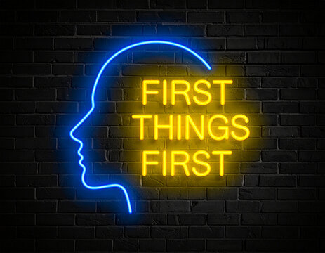 Leuchtende gelbe Neonschrift "First things first" umramt von einem blauen Neongesicht auf dunkler Backsteinmauer