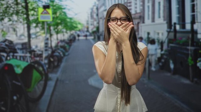 Teenage girl covers mouth with both hands on street, wearing glasses and white blouse; surprise candid moment.