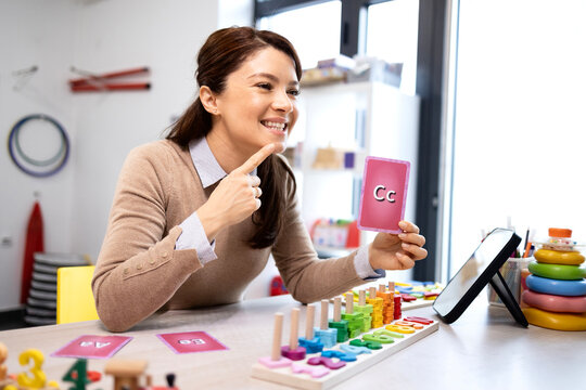 Female speech therapist on a video call holding an alphabet flashcard c while demonstrating mouth articulation for a child's remote phonics and language development lesson