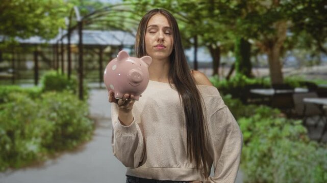Young woman holding pink piggy bank at arm's length on a street beside a park arbor and outdoor cafe tables; financial concern.