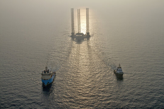 Netherlands, Netherlands - 13 February 2026: Aerial view of a towering oil rig piercing the misty horizon, flanked by two tugboats navigating the tranquil, sun-kissed waters.
