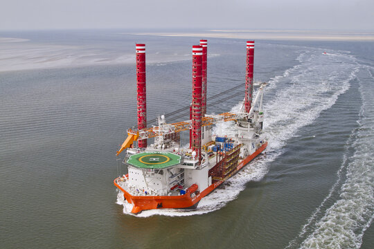 Esbjerg, Denmark - 15 February 2026: Aerial view of a vibrant orange and white oil rig vessel slicing through the grey-blue sea, leaving foamy white trails in its wake.