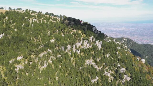 Panoramic drone shot of Spilios Agapitos refuge nestled among fir trees in the rocky landscape of Mount Olympus under a blue sky with clouds