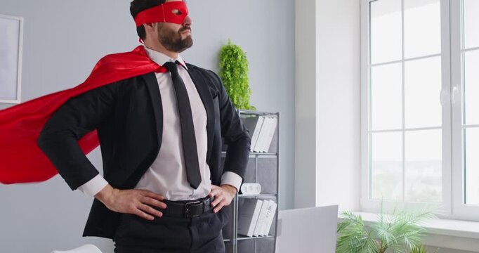 Portrait of young funny business man in superhero face mask and cloak working in office typing on a laptop. Confident cheerful male company employee standing on his workplace. 4k video.