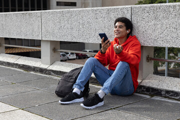 Man sitting on walkway by concrete barrier holding phone and wearing earbuds backpack, copy space © wavebreak3