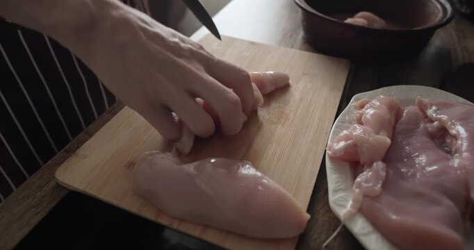 Close up of a professional chef slicing fresh raw chicken breast fillet on a wooden cutting board in a home kitchen, preparing healthy protein meat for cooking delicious dinner meals.
