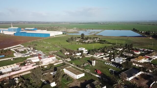 Drone shot capturing a strategic water storage basin for irrigation. Features the surrounding rural village, a modern industrial agriculture plant, and vast fertile fields in Northern Morocco.