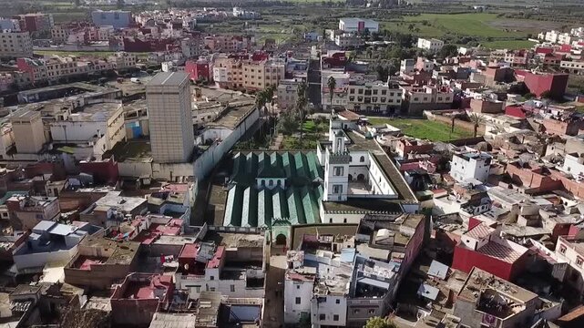 Captivating drone shot over the heart of Larache. Features the iconic green-tiled roof of the Grand Mosque, its white minaret, and the vibrant, dense urban architecture of the historic city.