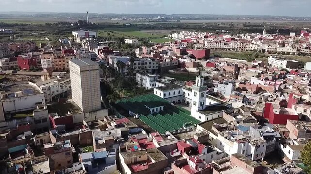 A detailed drone perspective of the historic Grand Mosque in Larache. Focuses on the elegant white minaret and the vast green-roofed prayer hall surrounded by the old city's urban texture.