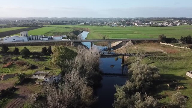 Dynamic drone shot over the irrigation canal in Larache. Features the contrast between modern and old bridges, a large industrial agricultural facility, and the surrounding green meadows.