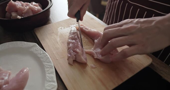 Close up of a professional chef slicing fresh raw chicken breast fillet on a wooden cutting board in a home kitchen, preparing healthy protein meat for cooking delicious dinner meals.